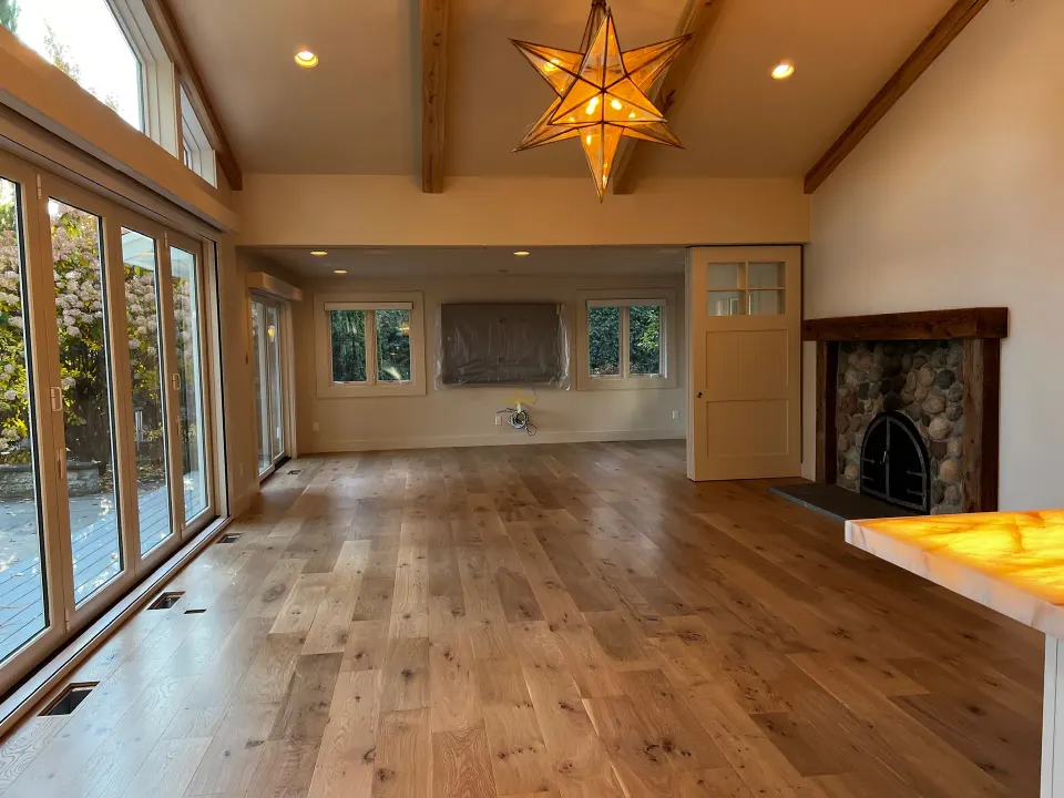 Great room with wide plank light oak flooring, a vaulted beamed ceiling, a wall of windows and a stone fireplace
