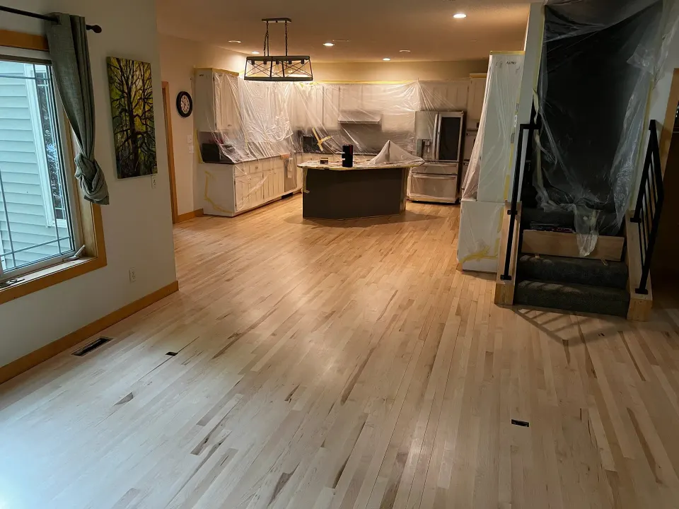 Maple floor sanded to bare wood partway through a refinish in a kitchen, with the cabinets and stairway masked in plastic