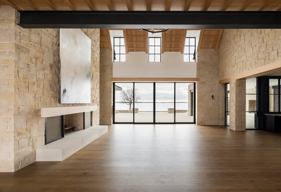 Great room with wide plank hardwood flooring, a stone fireplace wall and an exposed steel beam, with tall windows facing the water.