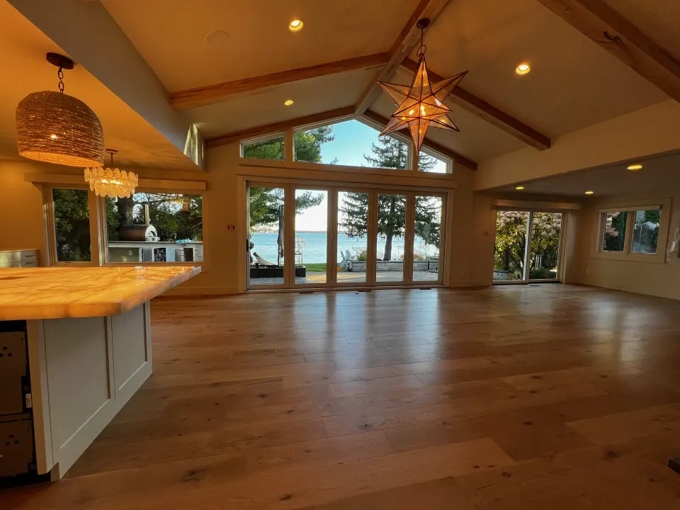 Vaulted great room with wide plank hardwood flooring and a wall of windows looking out over a lake.